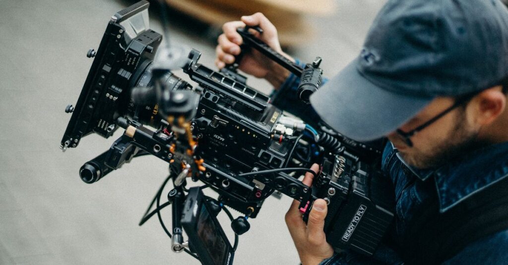 A man with a cap operates a professional video camera outdoors during daytime, capturing cinematic footage.