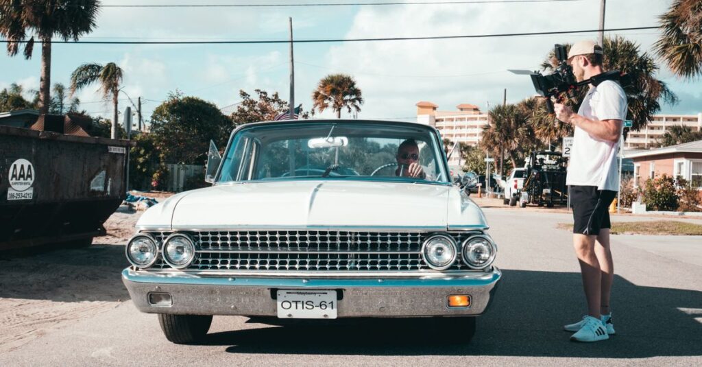 Filmmaker with handheld camera capturing a vintage car in a sunny urban street setting.