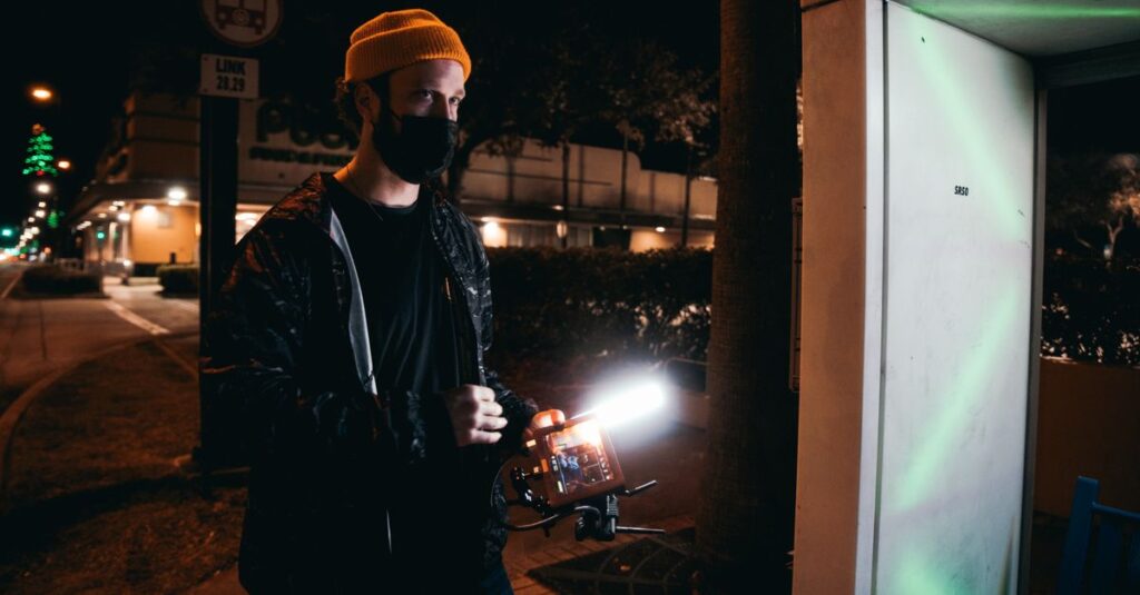 Man with camera gear capturing urban night scene, wearing a beanie and facemask.