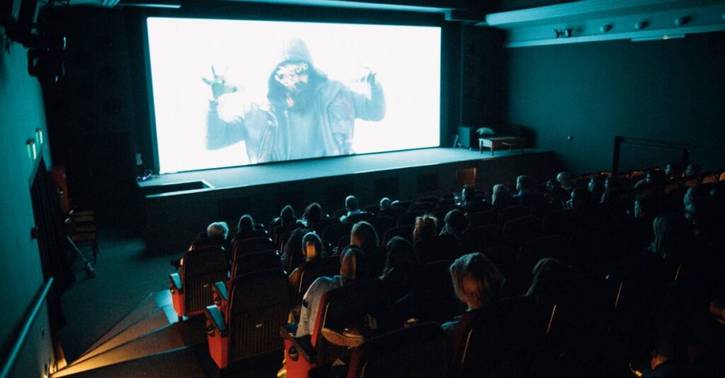 Crowded cinema theater with audience watching a movie on a large screen in a dark setting.
