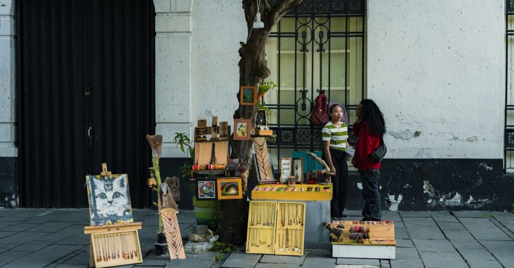 Two people converse at a street vendor stall displaying art and souvenirs on a city sidewalk.