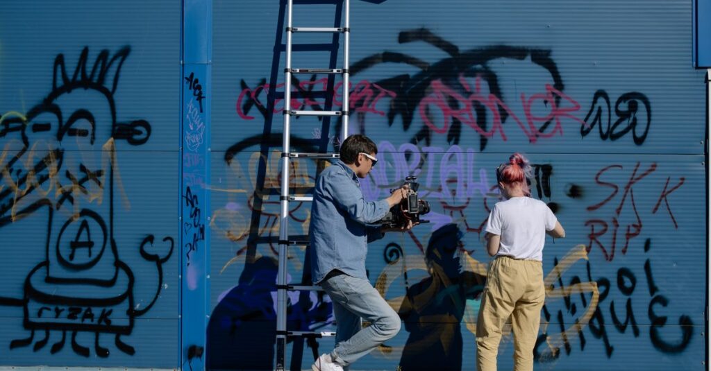 A man films a woman spray painting graffiti on a blue urban wall, showcasing street art.