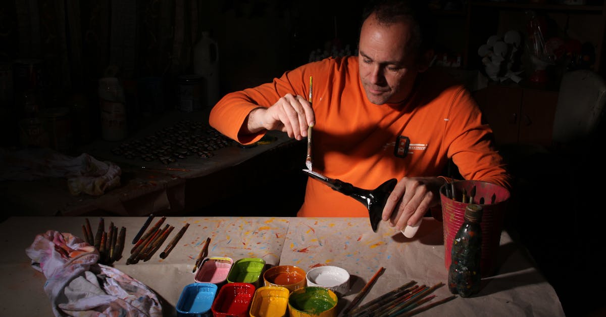 An artist in an orange shirt paints a model in a dimly lit studio, surrounded by colors and brushes.