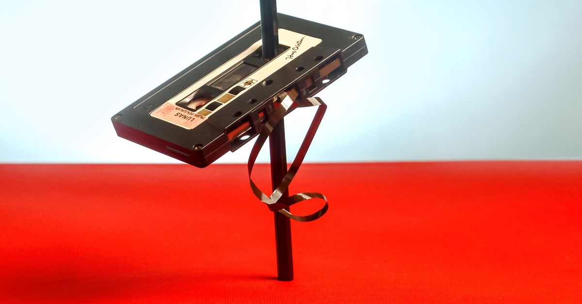 Creative still life of a cassette tape balanced with a pencil, vivid red background.