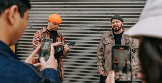 Street performers playing music while being filmed by a small crowd with smartphones.