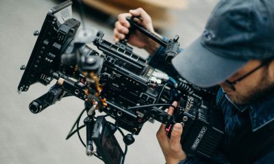 A man with a cap operates a professional video camera outdoors during daytime, capturing cinematic footage.