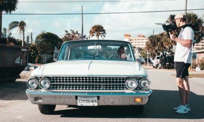Filmmaker with handheld camera capturing a vintage car in a sunny urban street setting.