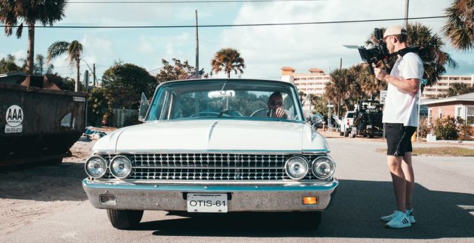 Filmmaker with handheld camera capturing a vintage car in a sunny urban street setting.