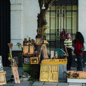Two people converse at a street vendor stall displaying art and souvenirs on a city sidewalk.