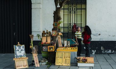 Two people converse at a street vendor stall displaying art and souvenirs on a city sidewalk.
