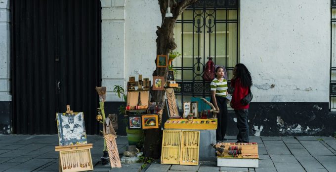 Two people converse at a street vendor stall displaying art and souvenirs on a city sidewalk.