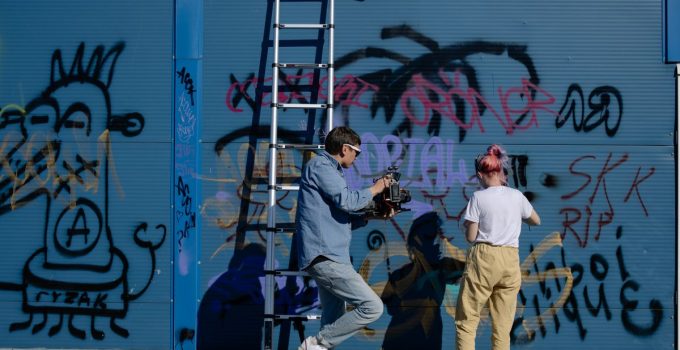 A man films a woman spray painting graffiti on a blue urban wall, showcasing street art.