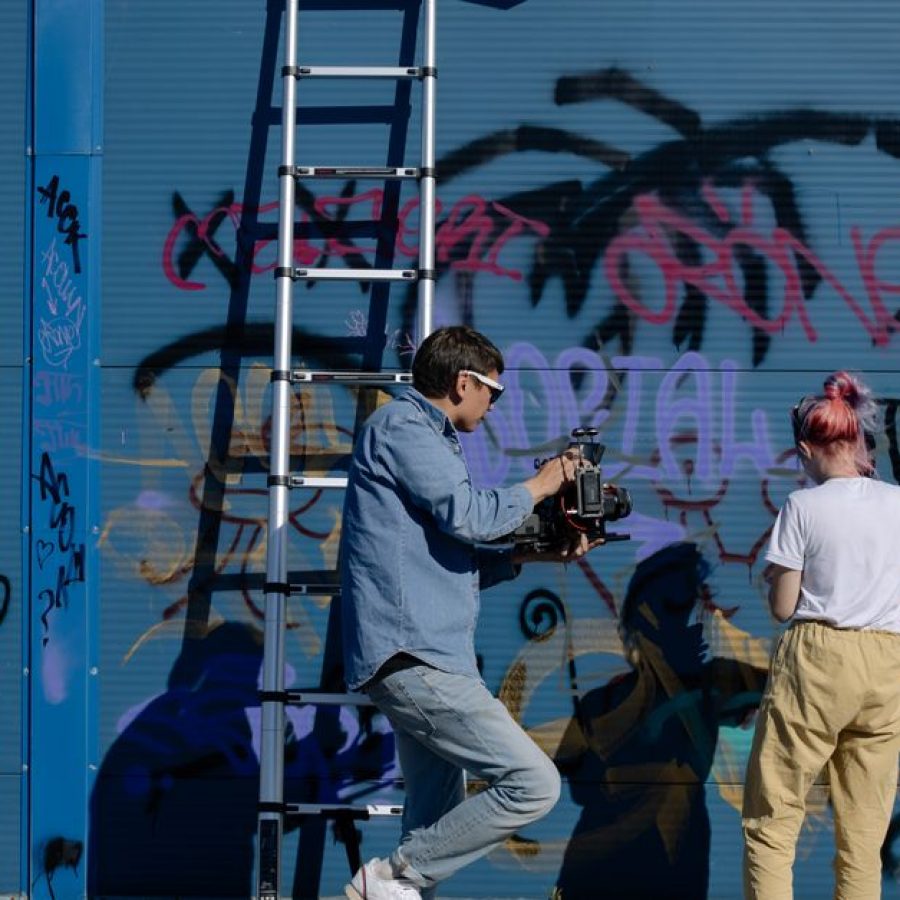 A man films a woman spray painting graffiti on a blue urban wall, showcasing street art.