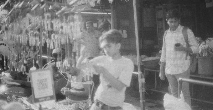 Artistic black and white photo capturing a bustling street market scene with people and hanging wares.